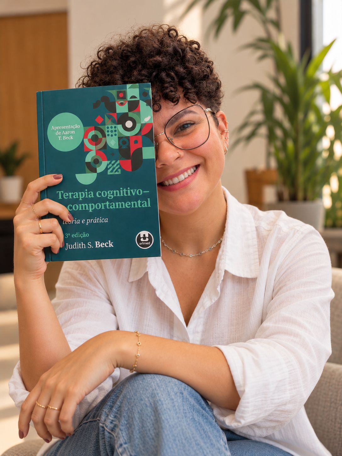 Poliana Almeida sorrindo, segurando ao lado do rosto o livro Terapia cognitivo-comportamental, de Judith S. Beck